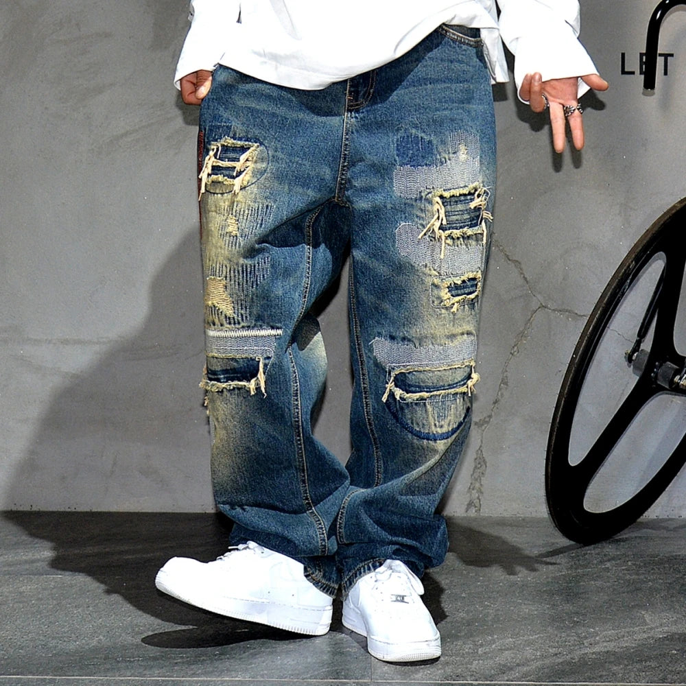 VOIDWEAR VDR_VANISH_05 ☰ baggy ripped patch jeans with distress wash, hip hop fit, ritual-coded glyph embroidery. Person wearing distressed blue jeans and white sneakers against a gray background.